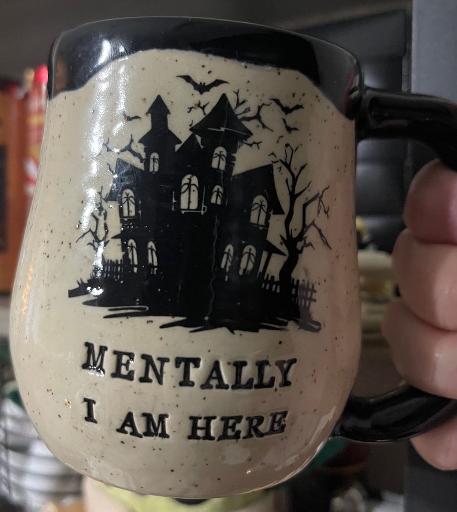 A handthrown mug that is cream with dark speckles, black seeping out over the rim with black ink artwork of an old victorian house that’s definitely haunted with soooky trees and bats flying around, below it reads “mentally I am here.”