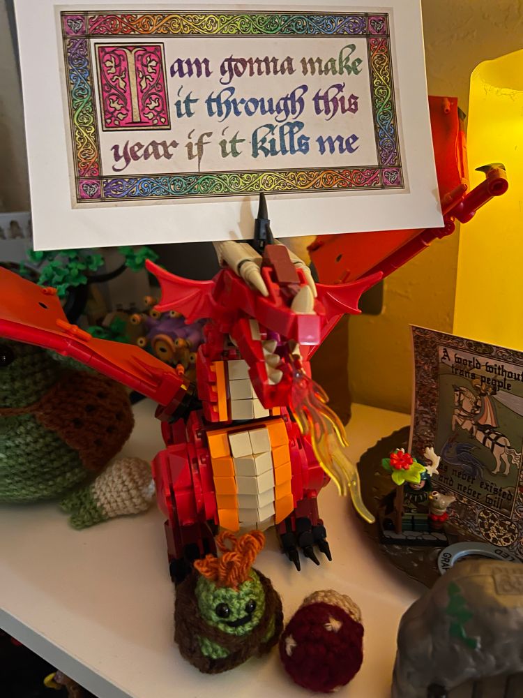 An art print sits above the head of a red and orange Lego dragon, from the DnD set. The print had a medieval frame around it in rainbow coloring. The text, also in medieval fonts and rainbow coloring reads “I am gonna make it through this year if it kills me” A small crochet goblin and mushroom and assorted things on the shelf. 

If you zoom in, you can see a smaller print from the same artist (link below and in reply to this), also medieval design, there’s a knight with the trans flag colors on their cloak, it’s putting a jousting lance through a dragon’s gullet. Text on it reads “A world without trans people has never existed and never will.” 

https://www.inprnt.com/gallery/outoutbriefcandle/