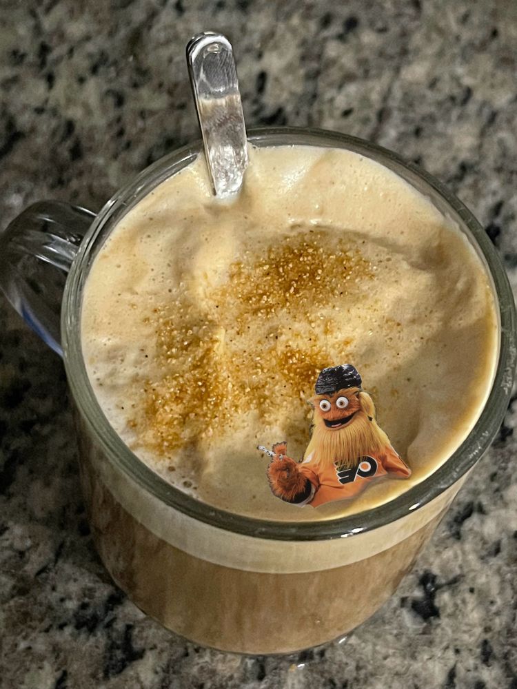 A glass mug of coffee with foam and brown sugar sprinkles on top. Atop the foam is Gritty, the Flyers mascot. 

Gritty is a furry orange monster with big eyes wearing a hockey helmet and jersey, long tuft of hair from head across the face right under the bottom lip. Gritty rules. 

It’s a sticker of a photo of Gritty I added to the pic of my coffee. 