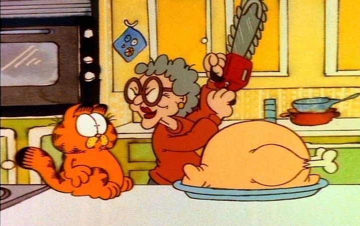 Still from the cartoon Garfield’s Thanksgiving. Garfield, a plump orange cat with black stripes is sitting on a counter, looking startled at an older woman with curly grey hair and big red rimmed glasses. She’s got a chainsaw in her hands, holding it up in the air. They’re in a kitchen, a large turkey sits on the counter in front of them. 