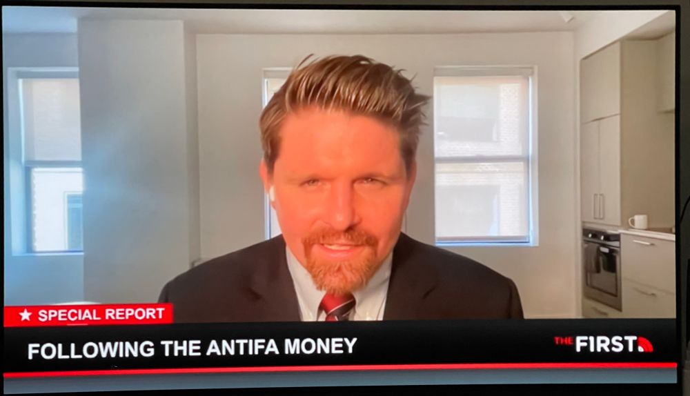 white guy with messy slicked back reddish blonde hair and beard, the lighting has his face looking very red. he’s in a suit. the room he’s in has white walls, 3 windows, there’s nothing on the walls, some of a kitchen can be seen. It has light gray cabinets. It doesn’t look like humans live there. 

The guy is Seamus Bruner.  