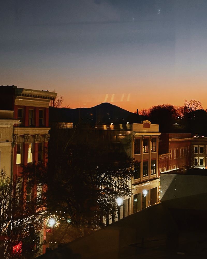 a sunrise featuring a historic city skyline and streetlights against a sky fading from dark blue to vivid orange. a silhouette of a mountain spindly trees is in the distance.