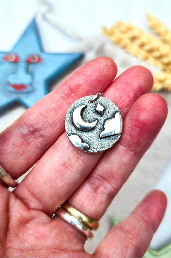 A handmade, round solid recycled silver pendant featuring a raised crescent Moon, a star, and two clouds against a textured background. The pendant is held between he maker's hand.