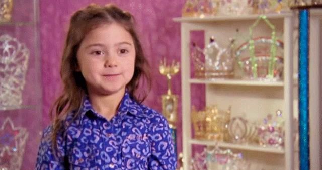 a little girl in a blue shirt is smiling in front of a display of tiaras .