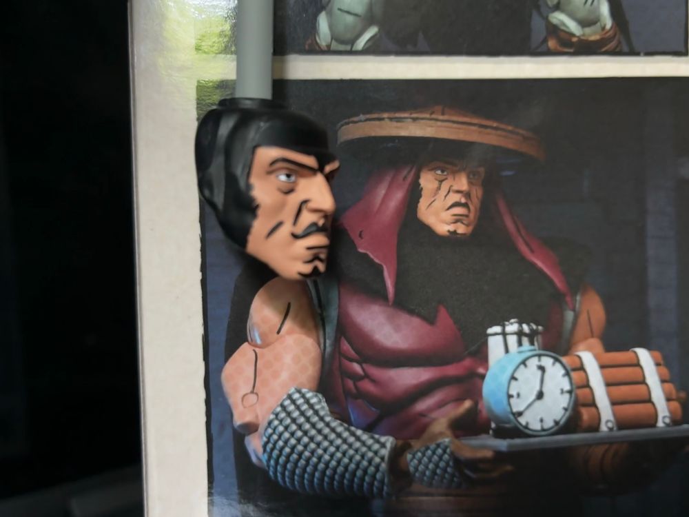 NECA TMNT Foot Elite Assassin action figure head compared with the picture of the same on the box. The box shows an upper lip shaded with black like a comic book might do while the finished product features a mustache instead.