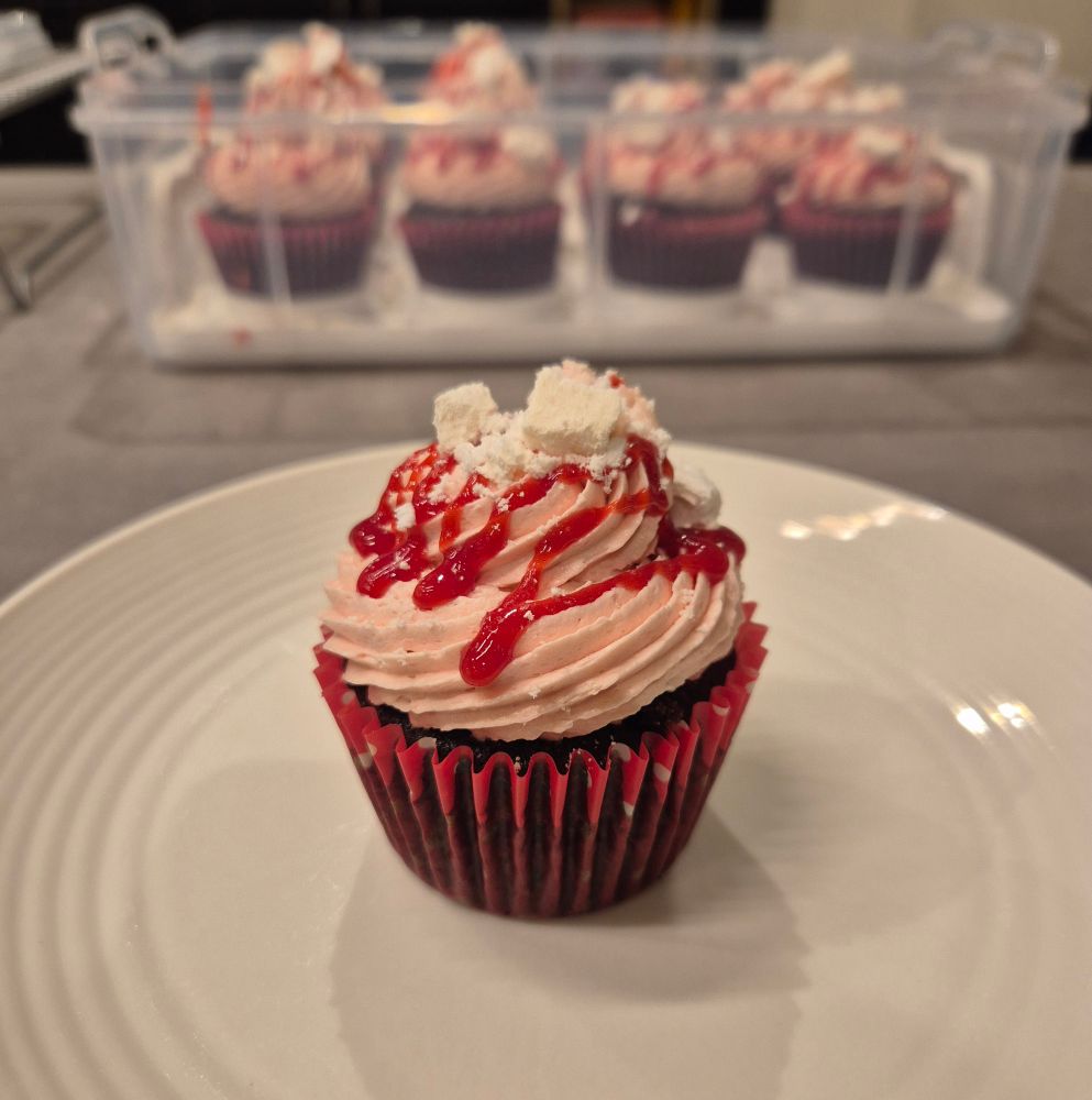 A single cupcake on a white side plate. The paper case is red with white polka dots and the cupcake itself is dark chocolate sponge (containing a hidden blob of homemade raspberry jam/sauce/goo) topped with a piped swirl raspberry buttercream and decorated with a drizzle of the raspberry goo and crushed French meringue. A clear plastic cupcake carrier holding the rest of the batch is out of focus in the background. 