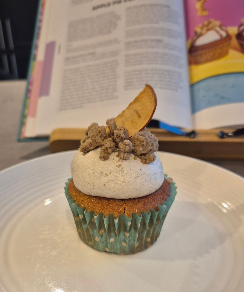 A single cupcake on a white side plate. It is an apple pie inspired cake and comprises a syrup sponge cake with an apple sauce filling, topped with a big splodge of cinnamon buttercream, and decorated with cinnamon pie crumbs (baked nuggets of cinnamon pastry) and a thin slice of apple baked to a crisp. It is delightful. 