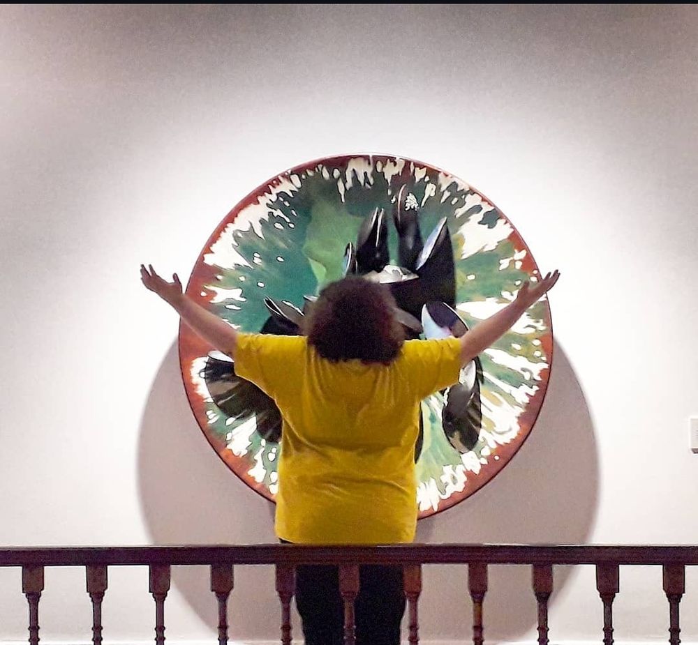 Estou no centro da imagem, um homem branco gordo, de camisa amarelo, cabelos loiros cacheados na altura dos ombros, de braços abertos, na frente de um painel redondo com vários formatos semelhantes a mexilhões, feitos por Adriana Varejão. Sobreposto, tem um corrimão de madeira de lei.