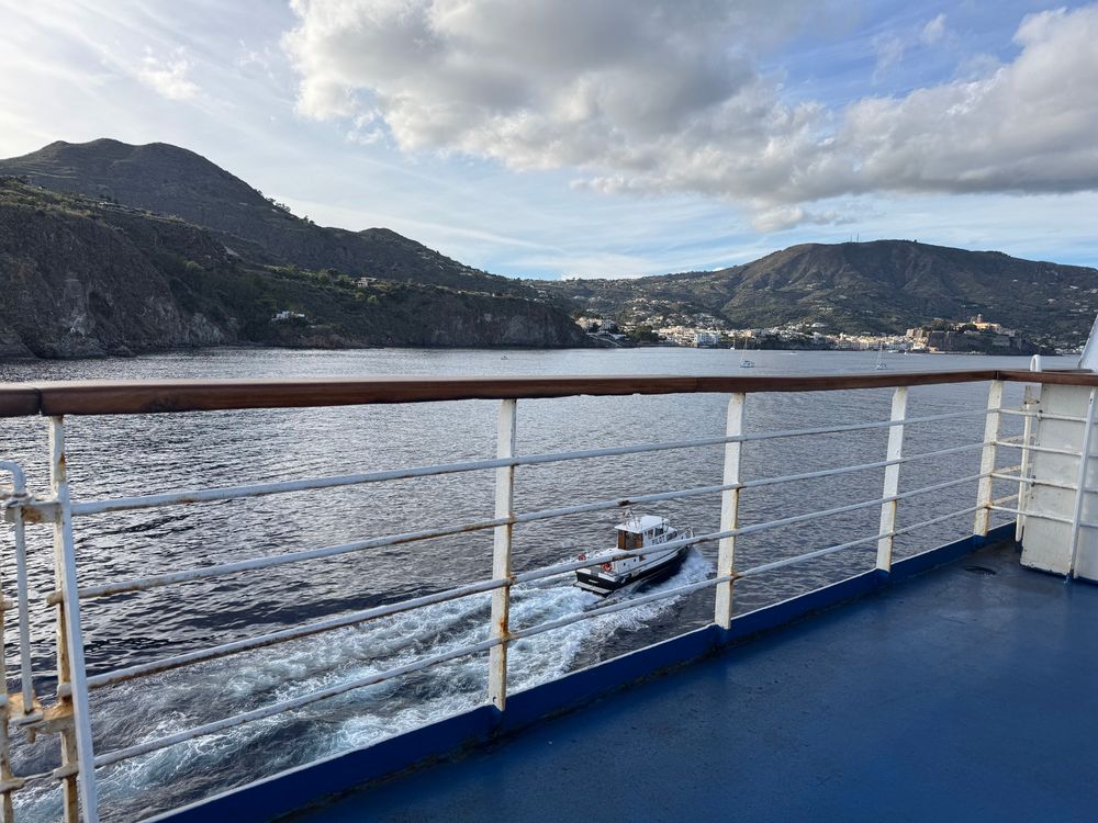 A small pilot boat sailing away between the ferry and the land. 