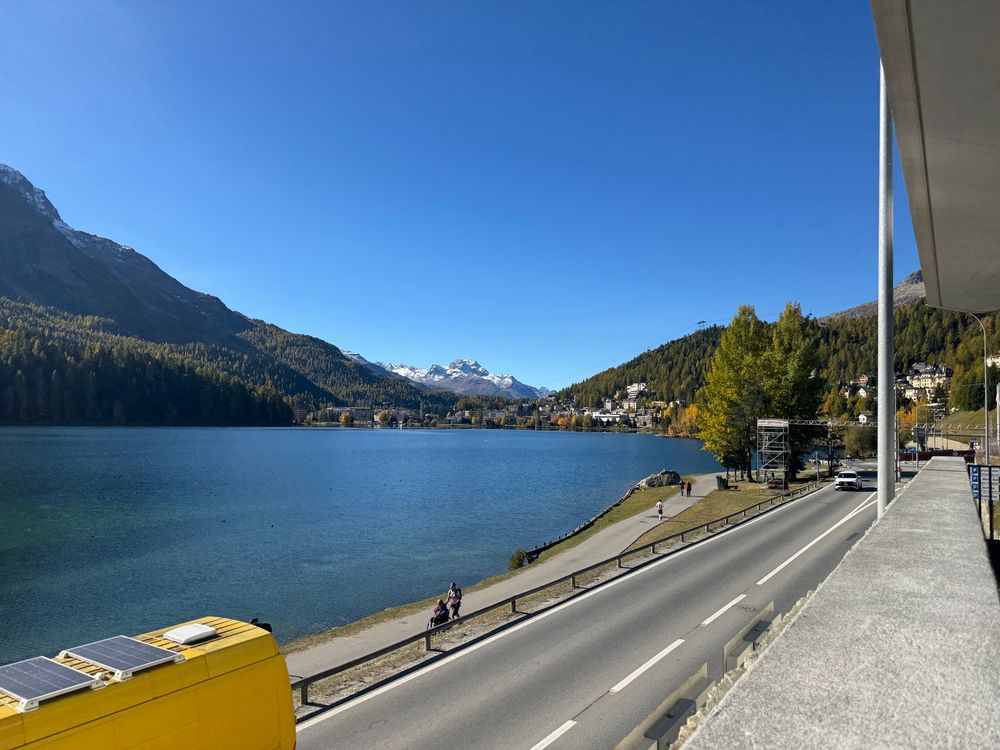 A lake ringed by mountains in sunshine. In the foreground is a road with a passing yellow van. 