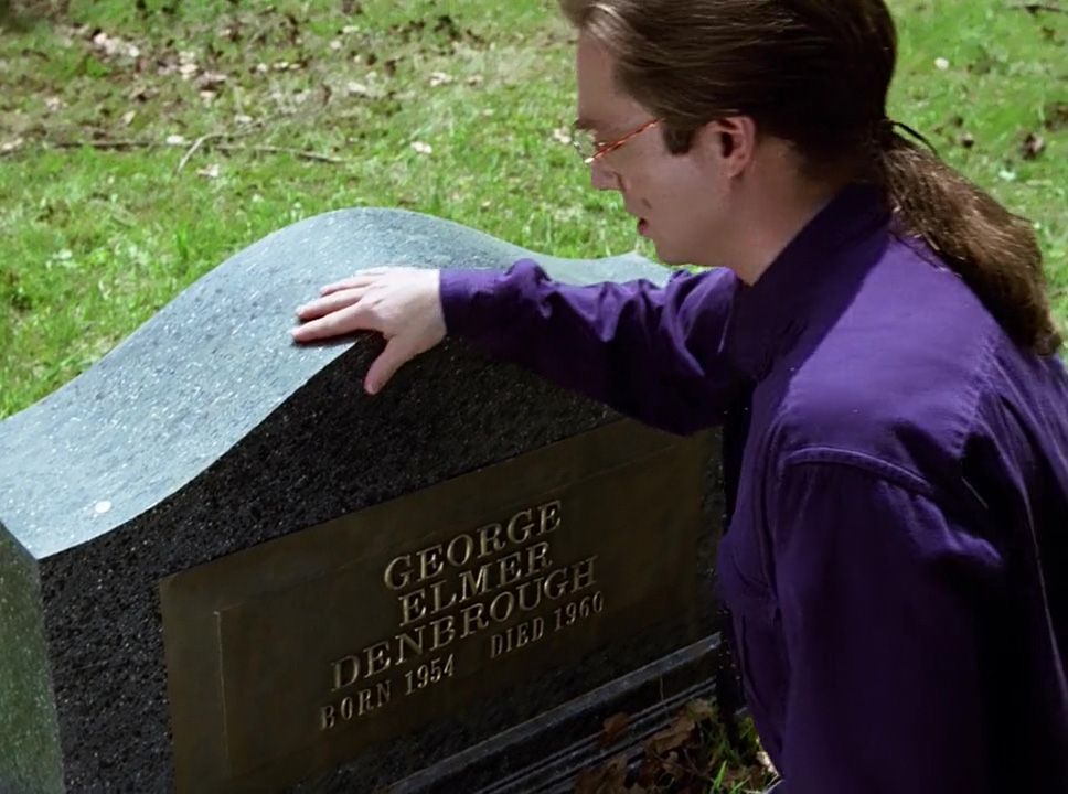 The character of Bill standing at the grave of his brother, wearing a purple shirt and sporting a long ponytail