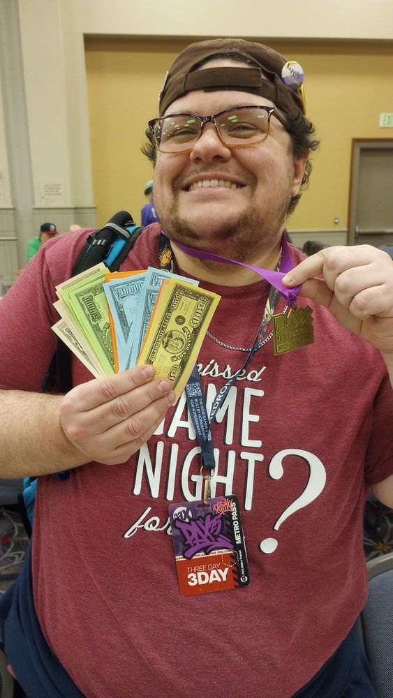 A grinning person holding play money and a 2025 PAX Unplugged gold medal.