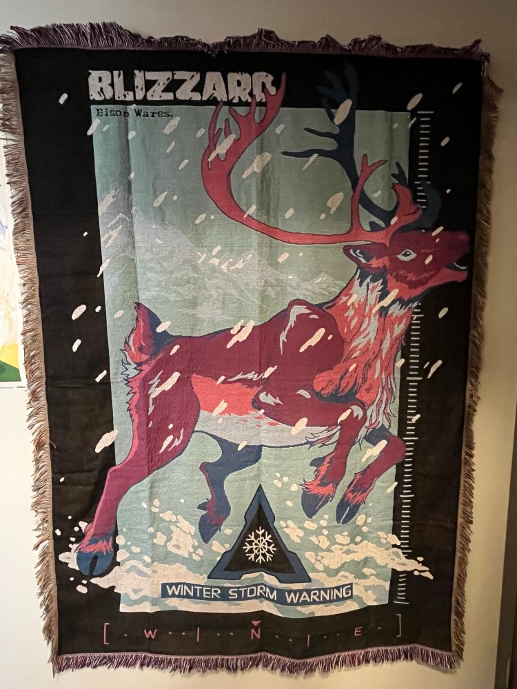 A photograph of a tapestry from Bisson wears of a caribou in a snow storm with the word blizzard on the top and compass like designs surrounding the image