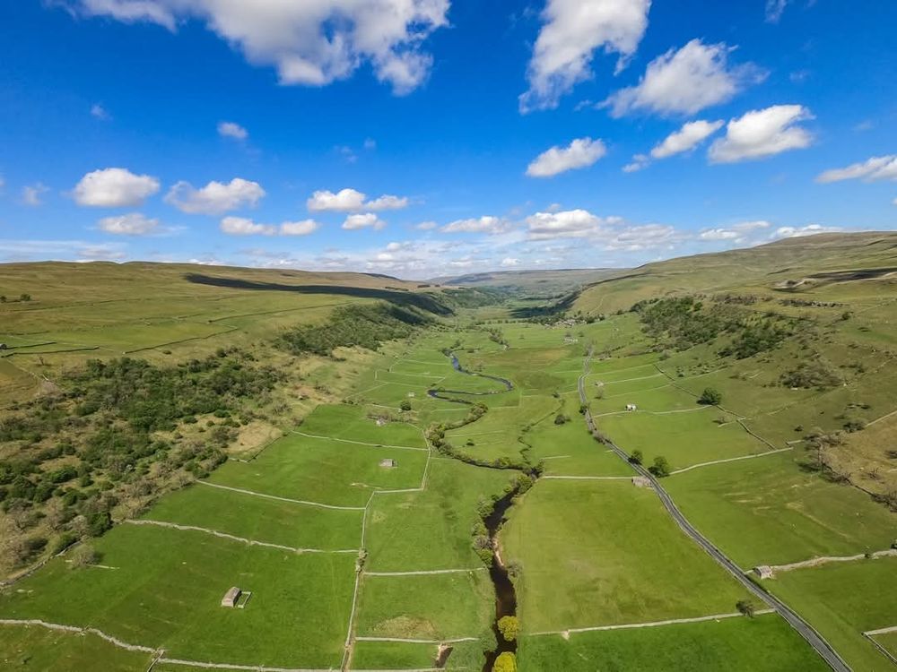 A river winds into a u shaped valley with bight green fields under a blue sky filled with fair weather clouds.