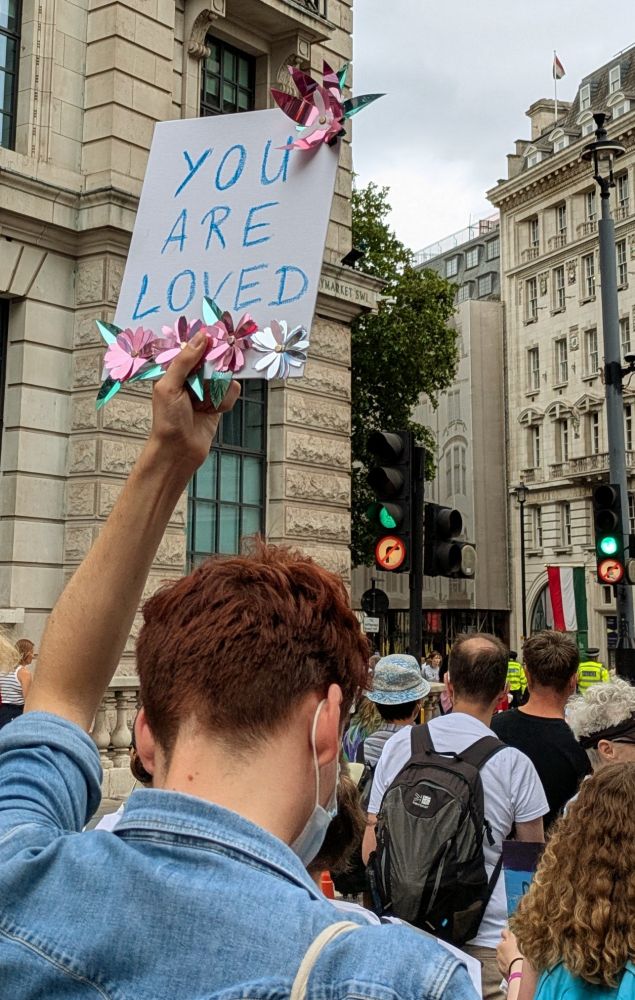 Person seen from behind on Trans Pride march holding up a simple sign saying YOU ARE LOVED 