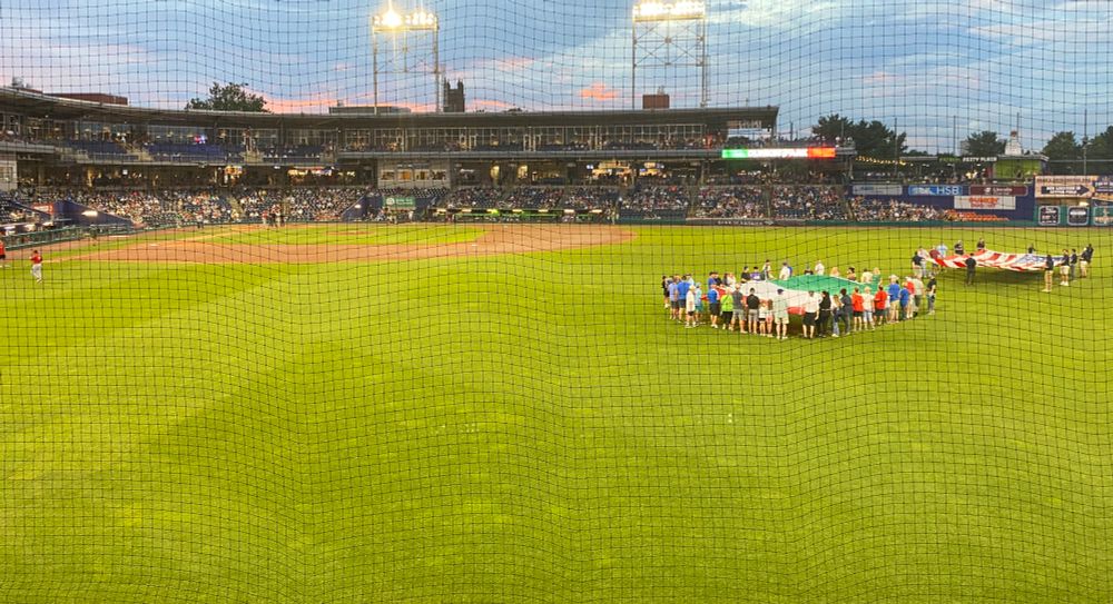 Picture of a baseball field at twilight (taken from the stands in right field looking in towards the diamond) with groups of people holding up a big Italian flag and a big American flag in shallow center field. 