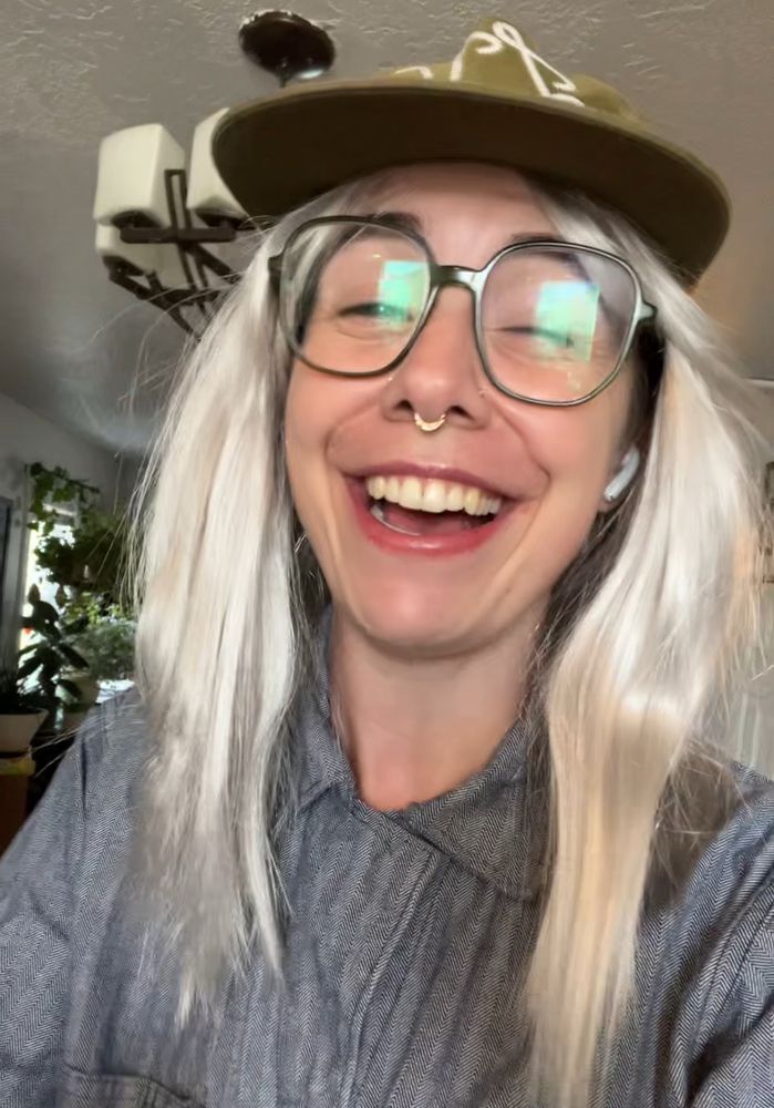 A photo of me smiling behind thick green rimmed glasses, a silver white long hair wig and a baseball hat 