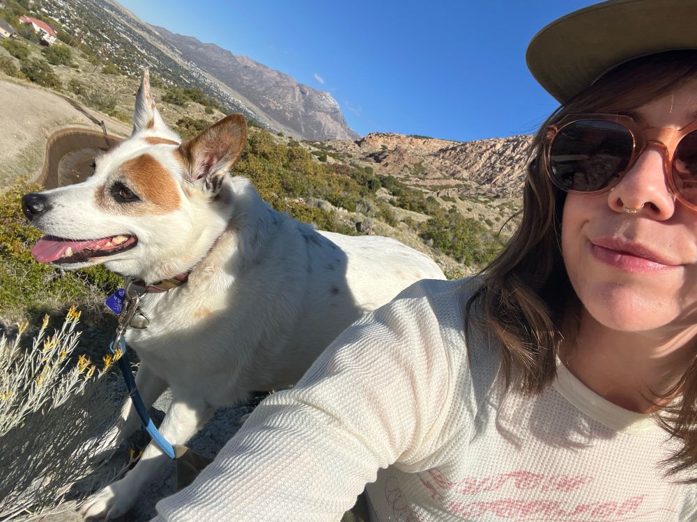 Me, a white person with brown shoulder length hair and sunglasses and a brown hat, sitting next to a white and brown heeler mix dog with a beautiful mountain behind us with green trees and brown steep cliffs 