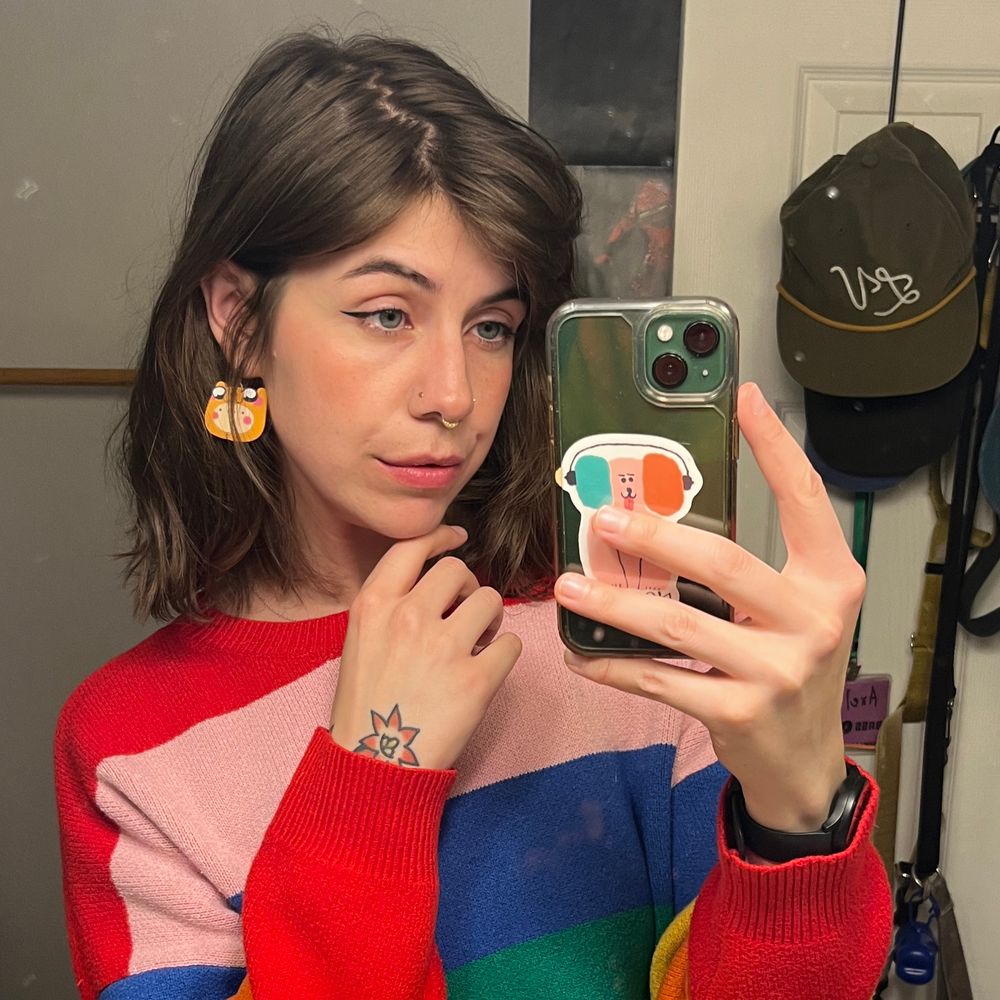 A mirror selfie of me - a white tomboy femme in a rainbow sweater, a Jake suit earring, and shoulder length brown hair 