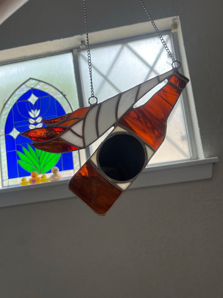Stained glass art of a brown bottle with a black label. There’s a white cloth strip on fire and it is coming out from inside the bottle. 