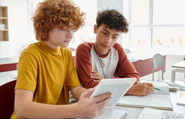 2 teenagers look at a tablet in a classroom setting. Photocredit LIGHTFIELD STUDIOS on Adobe Stock
