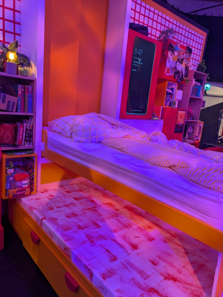 A theatre set containing a murphy bed, a screen in the wall, bookshelves and cabinets. The set is multicoloured and there is purple lighting on it.