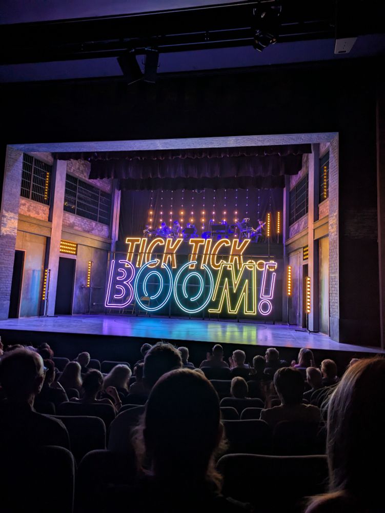 An audience in a theatre facing the stage. There is a band on the top level of the stage, and neon lighting that says "Tick Tick... Boom!"