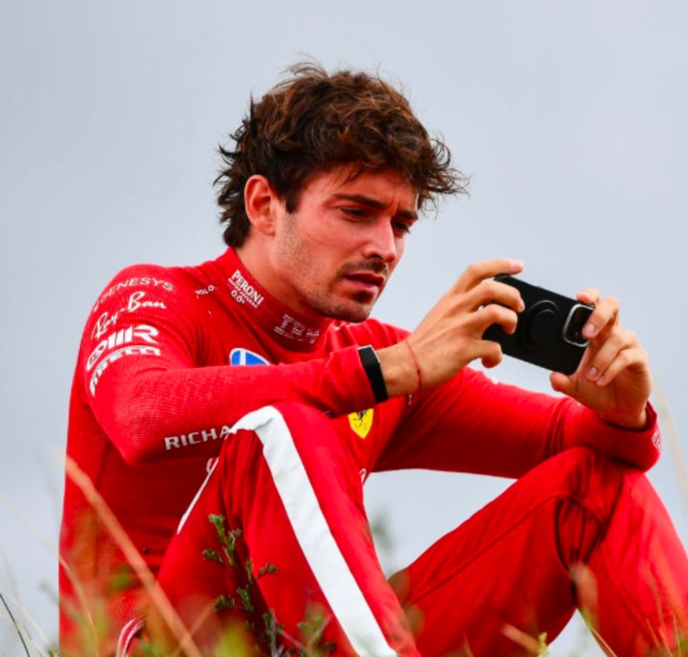 Charles Leclerc sat on grass and looking distressed while he watches something on a phone.
