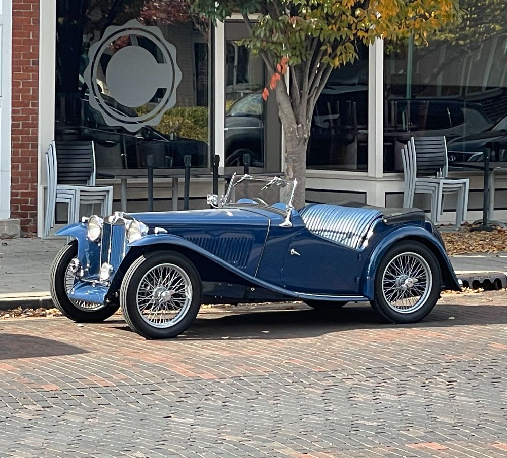 A blue MG TC parked on a brick street near a restaurant window emblazoned with a large C