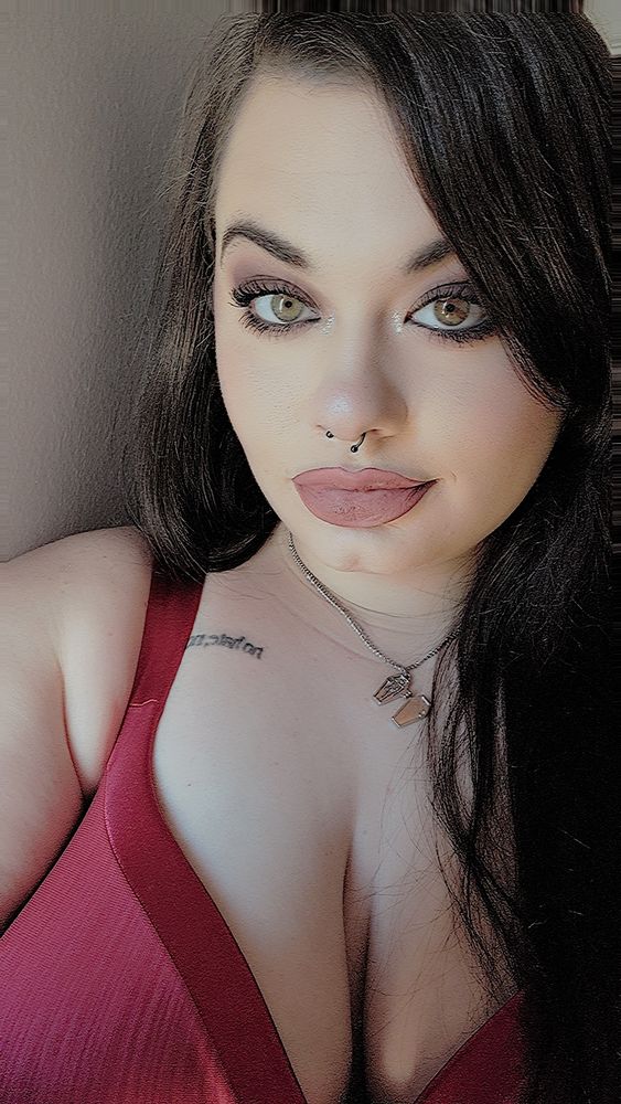 A close-up portrait of Alex, with long dark hair, light green eyes, and winged eye makeup. They have a septum piercing and wear a mauve lipstick. Alex is wearing a red top and a silver necklace with a small coffin charm. Soft natural light highlights their face and upper chest. 