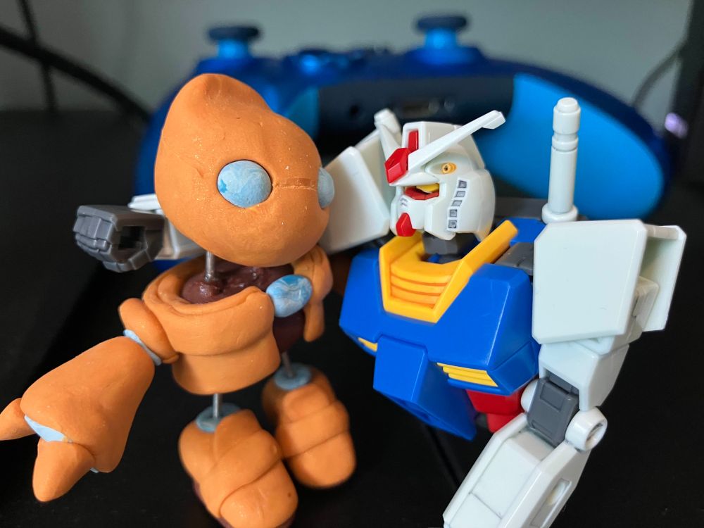 A clay model of Otto from AutoForge and a Gundam model stand side-by-side with their arms around each other.