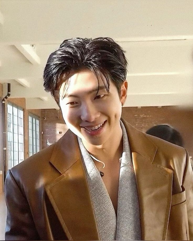 Kim Namjoon smiling, with black hair, wearing a brown overcoat in a salon  