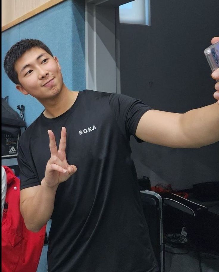 Namjoon in a black Korean army shirt, with his arm raised in a peace sign, a closed smile and a shaved head. his other hand holds the cell phone and he takes a picture. 