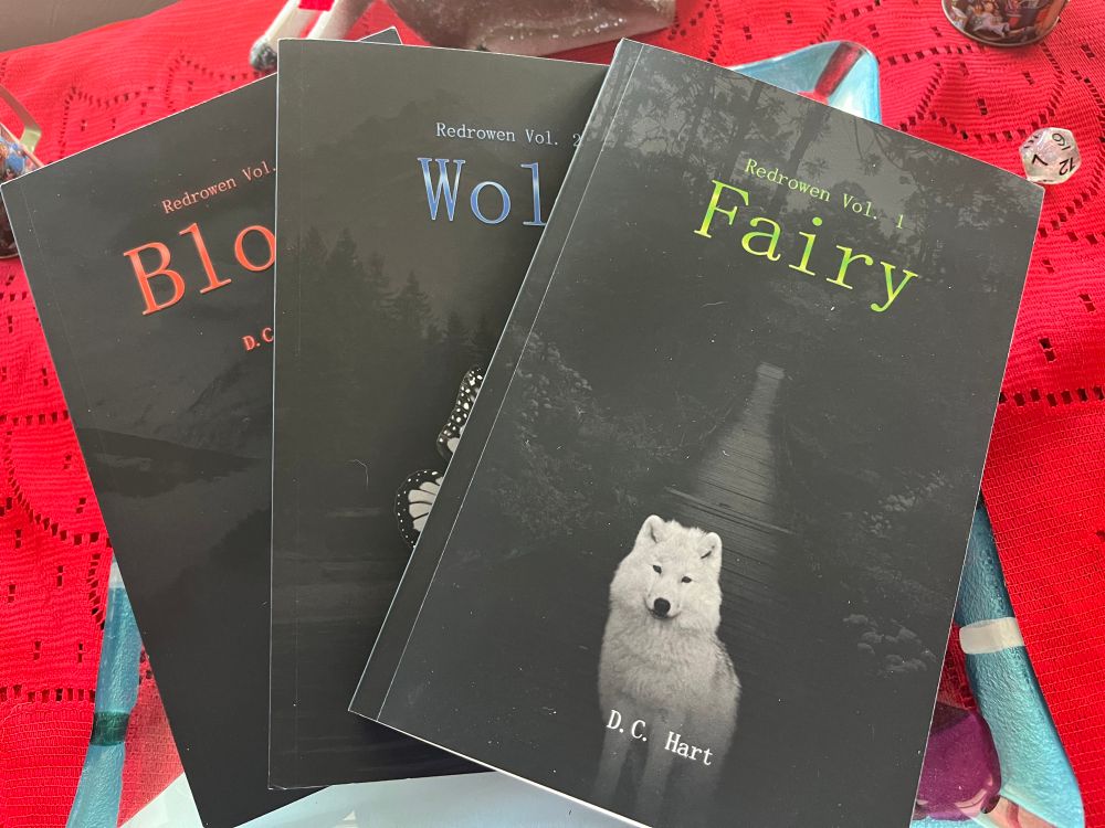 My books laid out against a red backdrop. The top book (my first) is Fairy. The cover features a wolf against a darkened background with green typeface. 