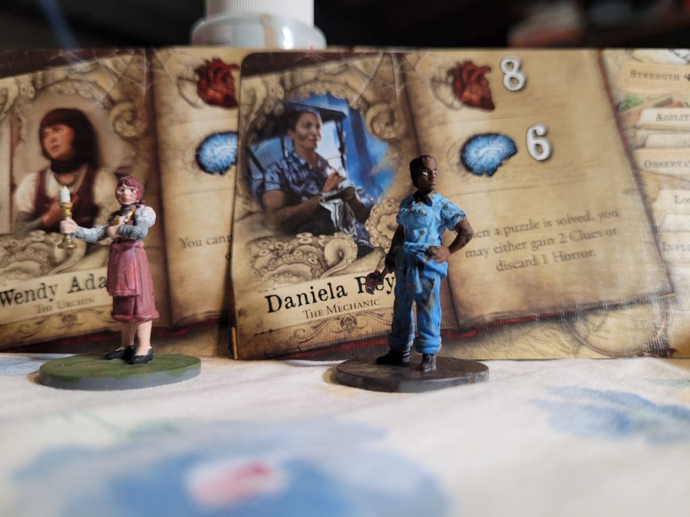 Photo of painted figures of Daniela and Wendy from the game mansions of madness. They are standing in front of their respective character cards 