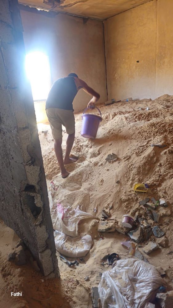 Fathi uses a bucket to clear sand out of his home. There whole area is buried in like 4 feet of sand. There is also old trash bags strewn in the foreground 