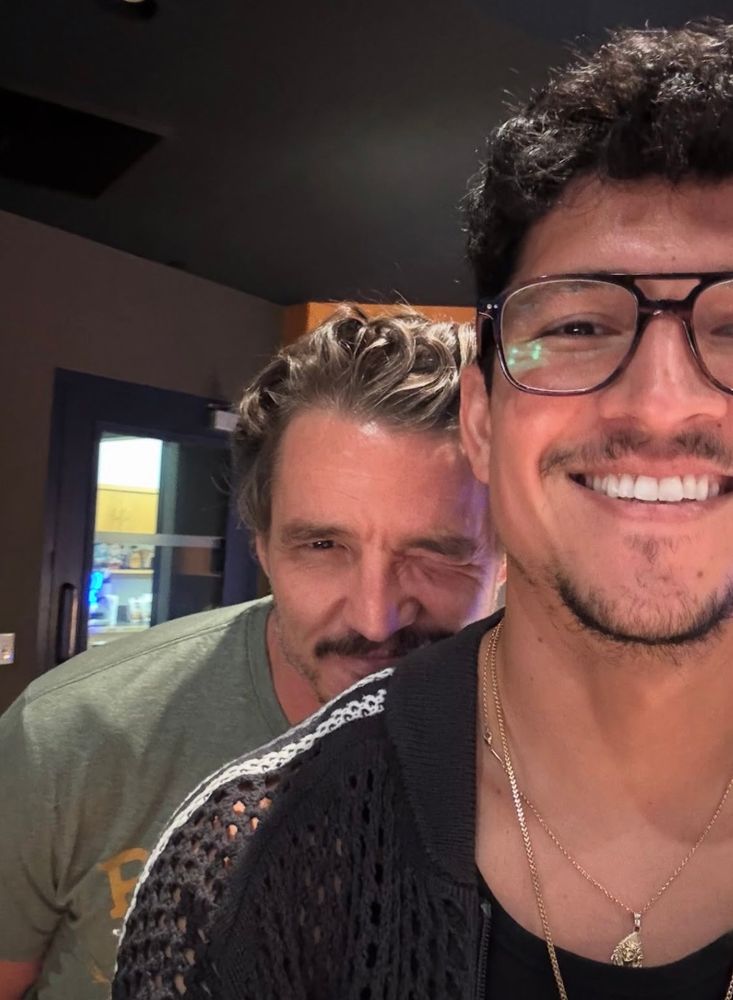 Photo showing Danny Ramirez wearing glasses and standing in the foreground with Pedro Pascal peeking over his shoulder.