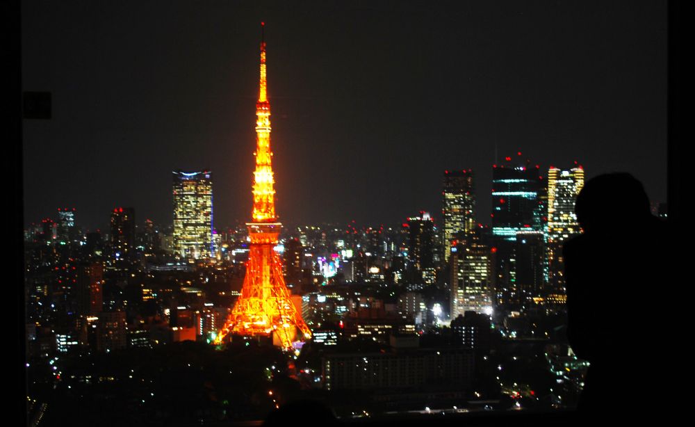 Night skyline of Tokyo with Tokyo Tower burning bright in the middle of it all. To the far right the silhouette of a man looking out of the window with this view.