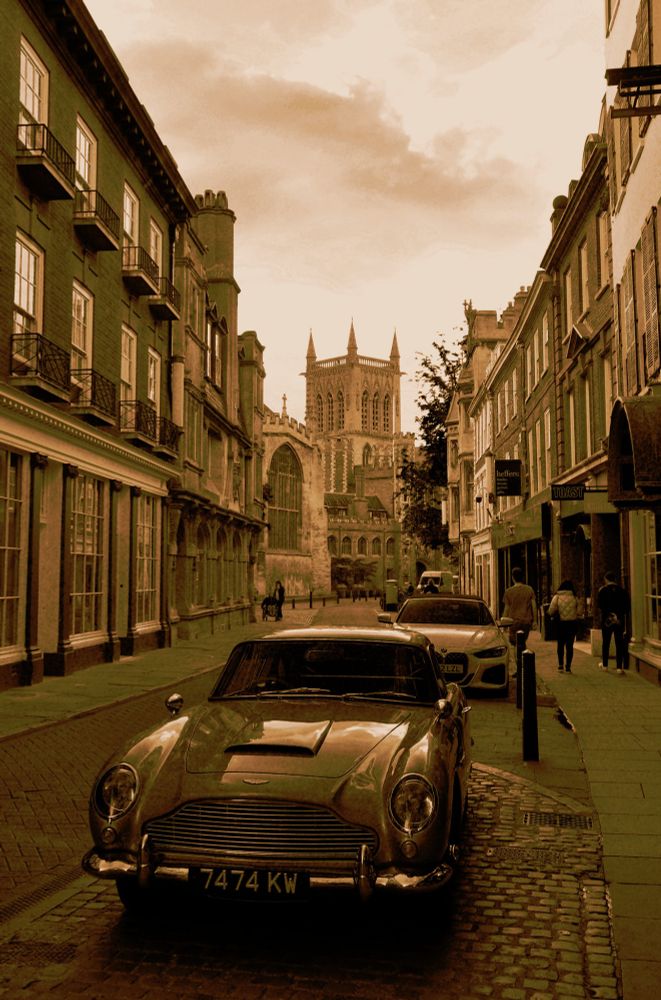 Sepia toned photo of a street with shops along the sides, and with a Gothic church in the background. In the centre of the photo is an Aston Martin D85 parked.