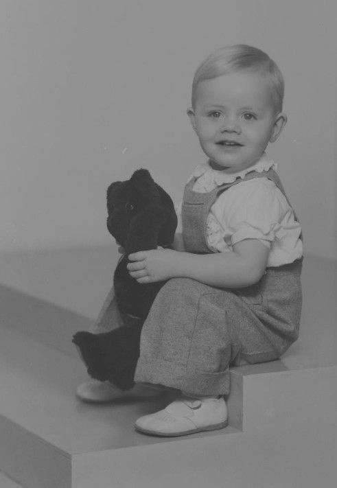 Portrait of a sitting boy, almost 2 years old, holding a teddybear in his hands, looking at the camera. He is blond, wearing trousers and a short-sleeved shirt, and is smiling at the camera. Photo taken in December of 1947.