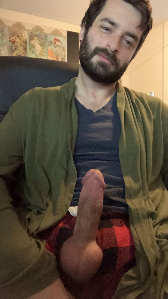 A bearded man in a cardigan and pajama pants facing the camera. His erect penis is coming through the fly of his pants 