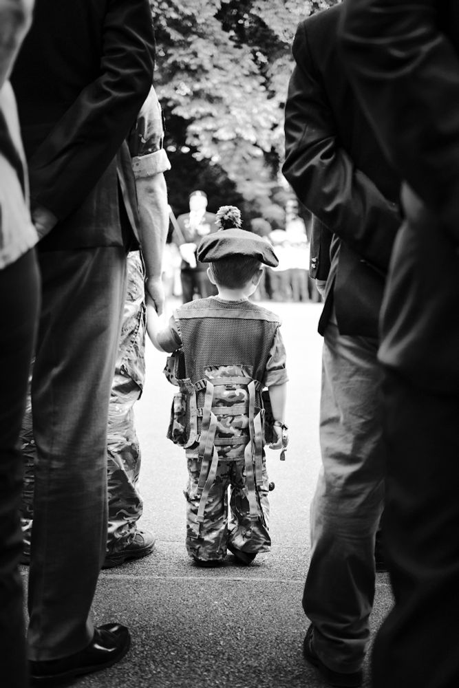 A small child dressed in an army combat uniform stands holding the hand of his soldier dad during an armed forces day parade