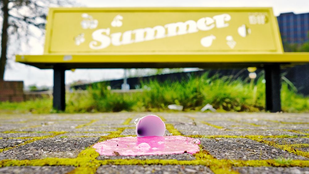 A melted pink icecream on the pavement, in front of a yellow bench bearing the word ‘summer’