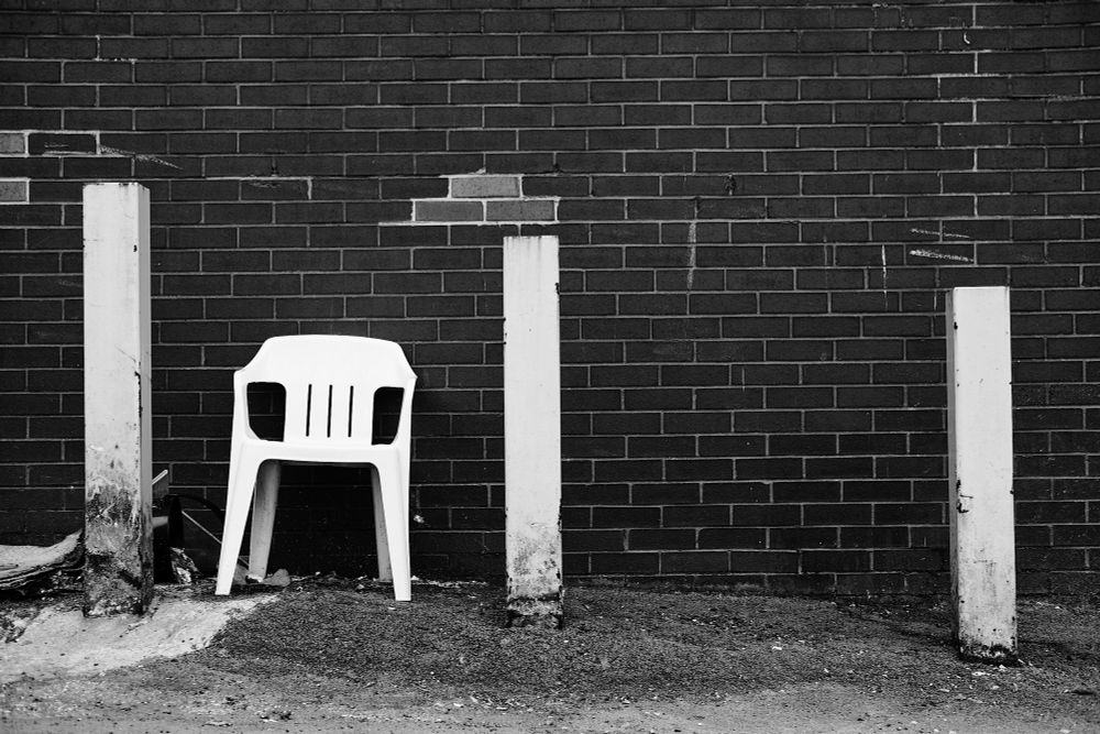 A white plastic chair between white metal parking bollards against a brick wall in space behind a shop
