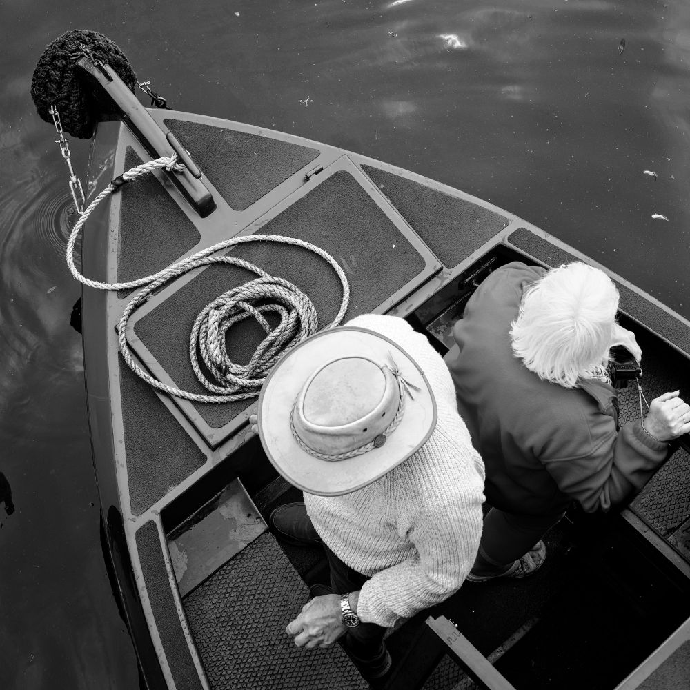 Viewed from a bridge, looking directly down, a man in a white hat, and a woman with white hair, manoeuvre their canal boat slowly along a canal. Black and white photograph