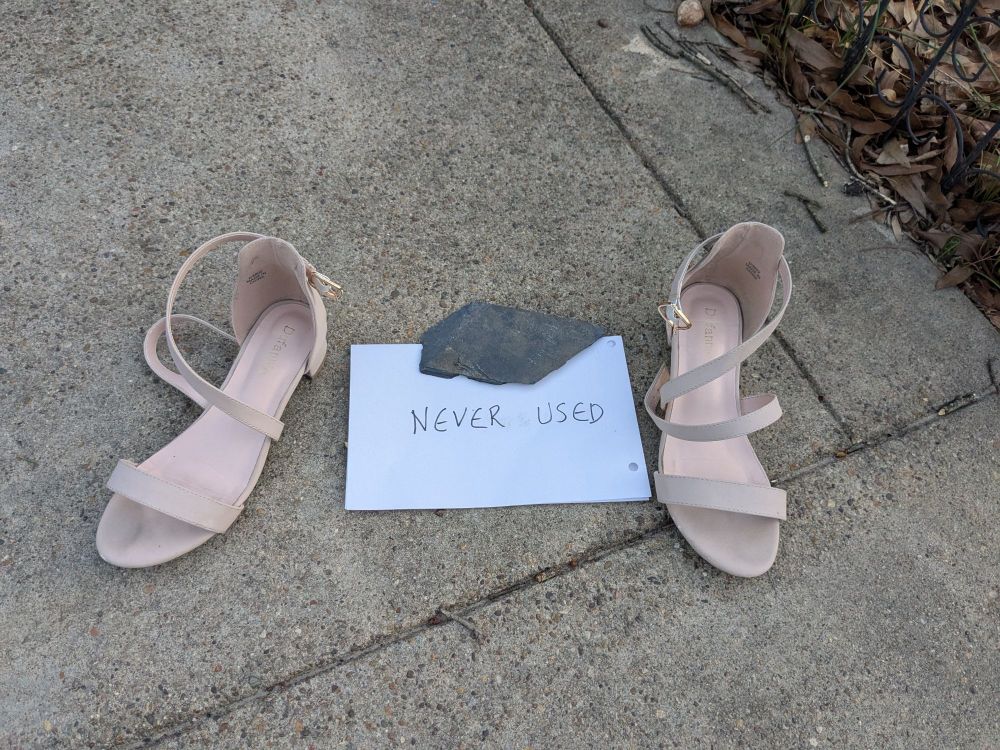 Pink heeled women's shoes on the sidewalk with a sign saying never used. Shoes look worn.