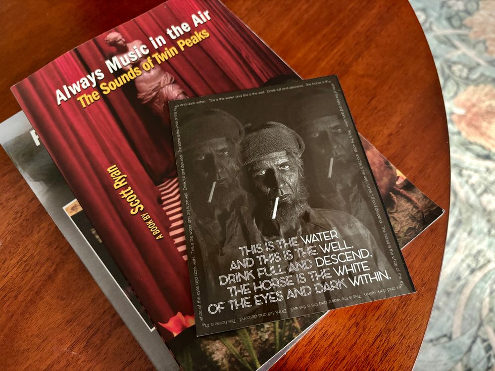 Two paperback books, one showing the title "Always Music In the Air: The Sounds of Twin Peaks," on top of them a large rectangular magnet with images of the Woodsman from TWIN PEAKS: THE RETURN Episode 8 and the quote: "This is the water and this is the well. Drink full and descend. The horse is the white of the eyes and dark within."