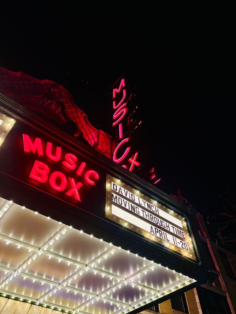 Against a black night sky, a portion of theater marquee glows as seen from underneath, with neon red letters “Music Box” and sign reading “David Lynch Moving Through Time April 11-20.”