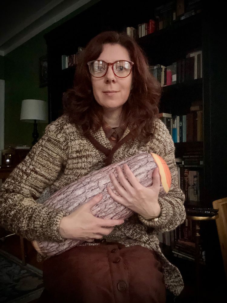 Me, a white woman with long brown hair, wearing a mottled brown sweater, brown skirt and red-rimmed eyeglasses, holding a pillow shaped like a log.