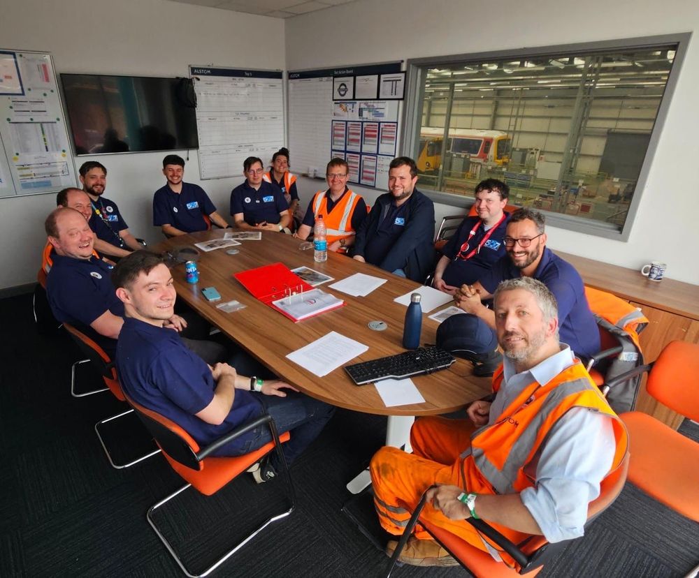 Photo of the C507PS team sitting around a conference table in an office, with trains visible out of the window in V Shop beyond 